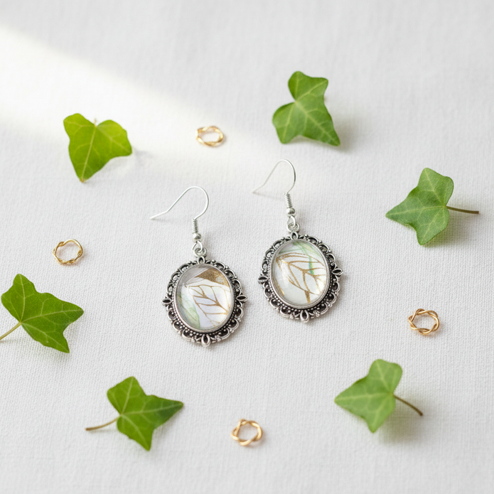 Botanical leaf cabochon drop earrings flat lay on white linen with green leaves and gold accents