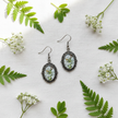 Small botanical cabochon drop earrings flat lay on white linen with green leaves and white flowers