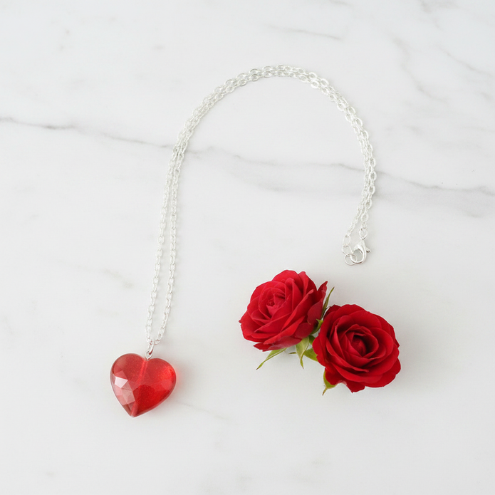 Faceted red glass heart pendant on silver chain necklace on marble surface with red roses