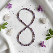 Flat lay of purple and brown beaded necklace with silver spacers on white linen with purple flowers