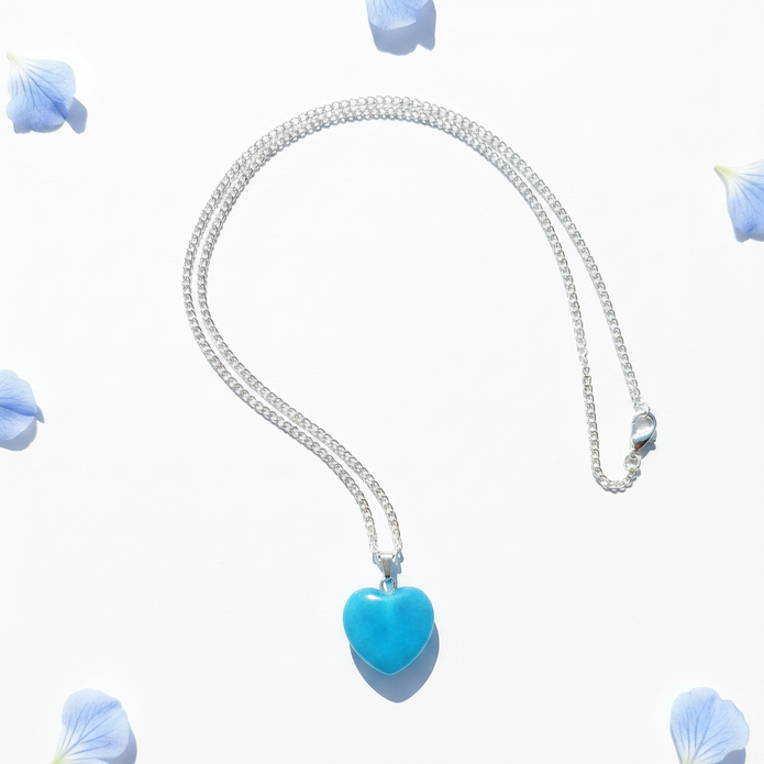 Turquoise blue glass heart pendant necklace flat lay with blue flower petals on white background