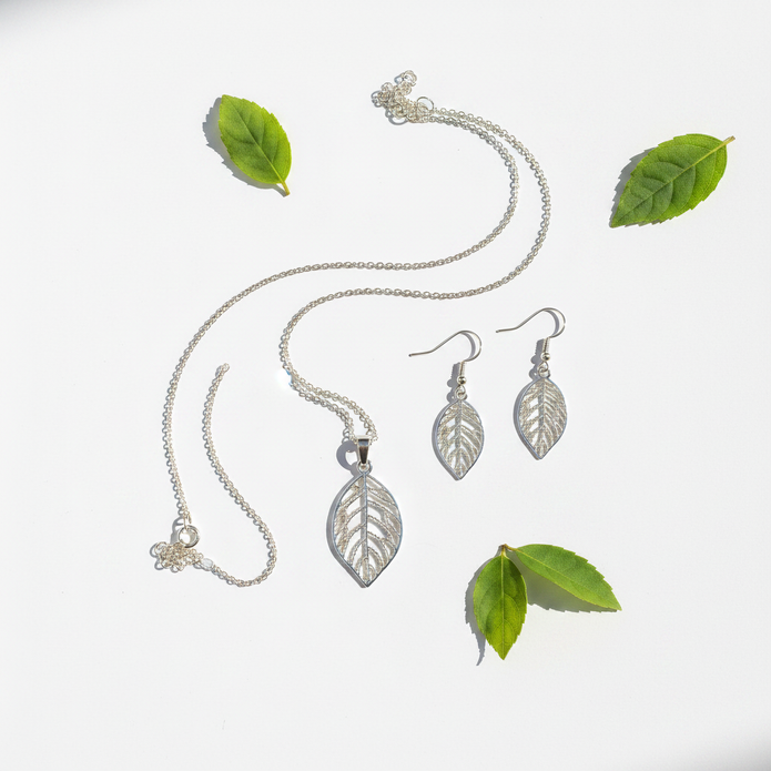 Silver filigree leaf necklace and earrings set flat lay with green leaves on white background
